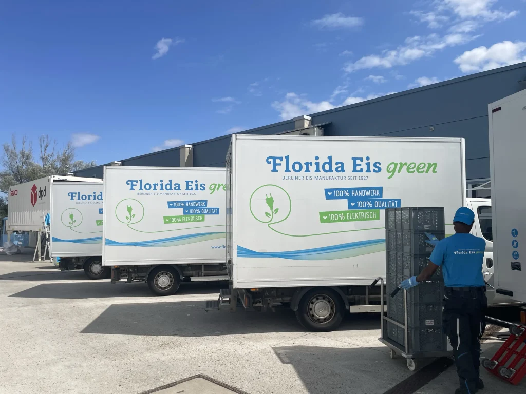 Zwei Lieferwagen von Florida Eis green stehen vor einer Lagerhalle unter blauem Himmel. Im Vordergrund schiebt ein Mitarbeiter in blauer Arbeitskleidung einen hohen Rollcontainer mit schwarzen Transportkisten. Auf den elektrisch betriebenen Lieferfahrzeug